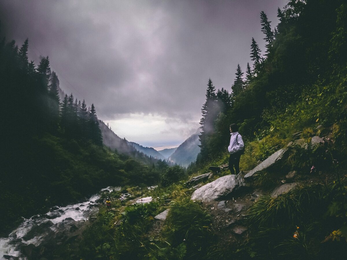 A person stands on a rocky trail beside a stream, surrounded by dense forest and mountains under a cloudy sky.