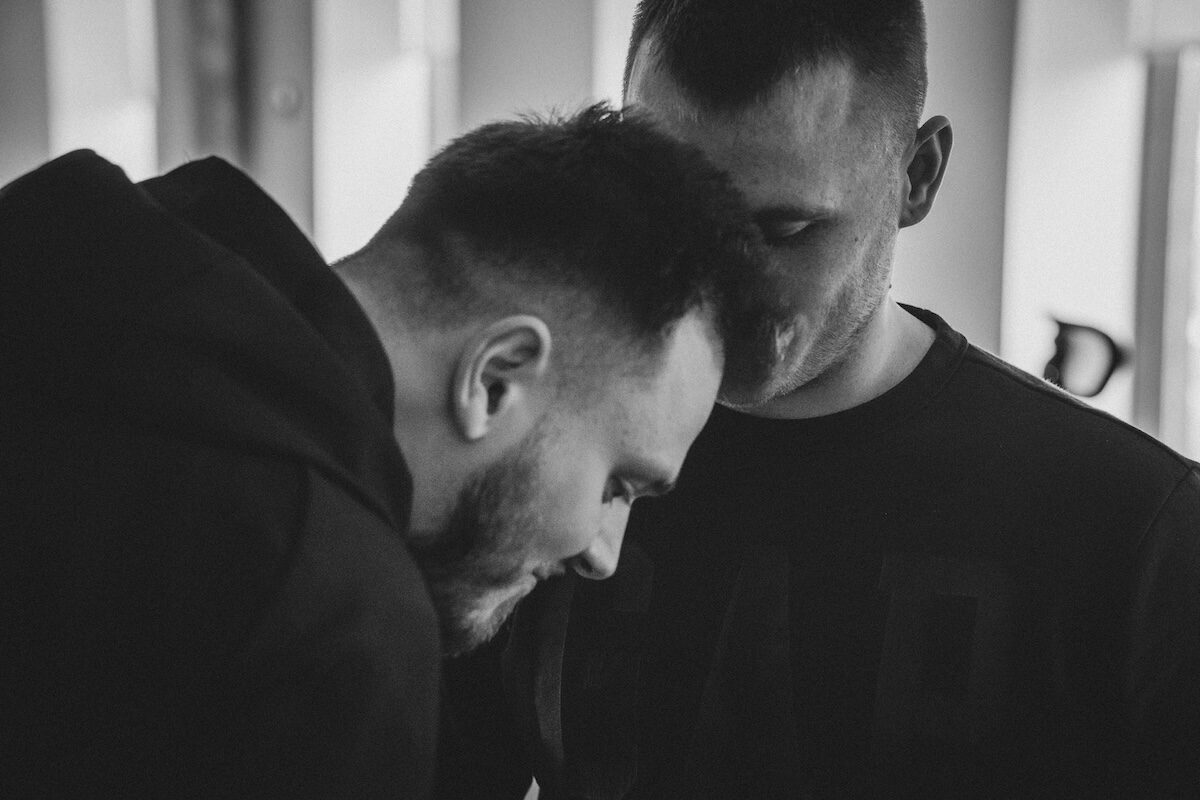 Two men are standing close together indoors, one leaning his head forward while the other looks down. The image is in black and white.