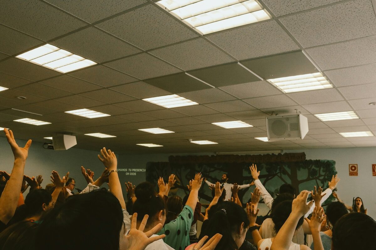 A group of people in a room with hands raised, facing forward under fluorescent ceiling lights.