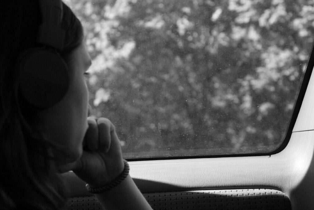 Person wearing headphones looks out a car window, resting their chin on their hand. Trees are visible outside in the background. The photo is in black and white.