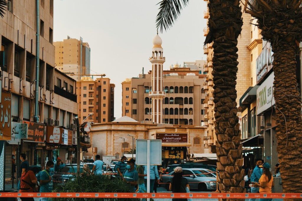 A busy city street with people walking, cars parked, palm trees, and a mosque with a tall minaret in the background surrounded by buildings.