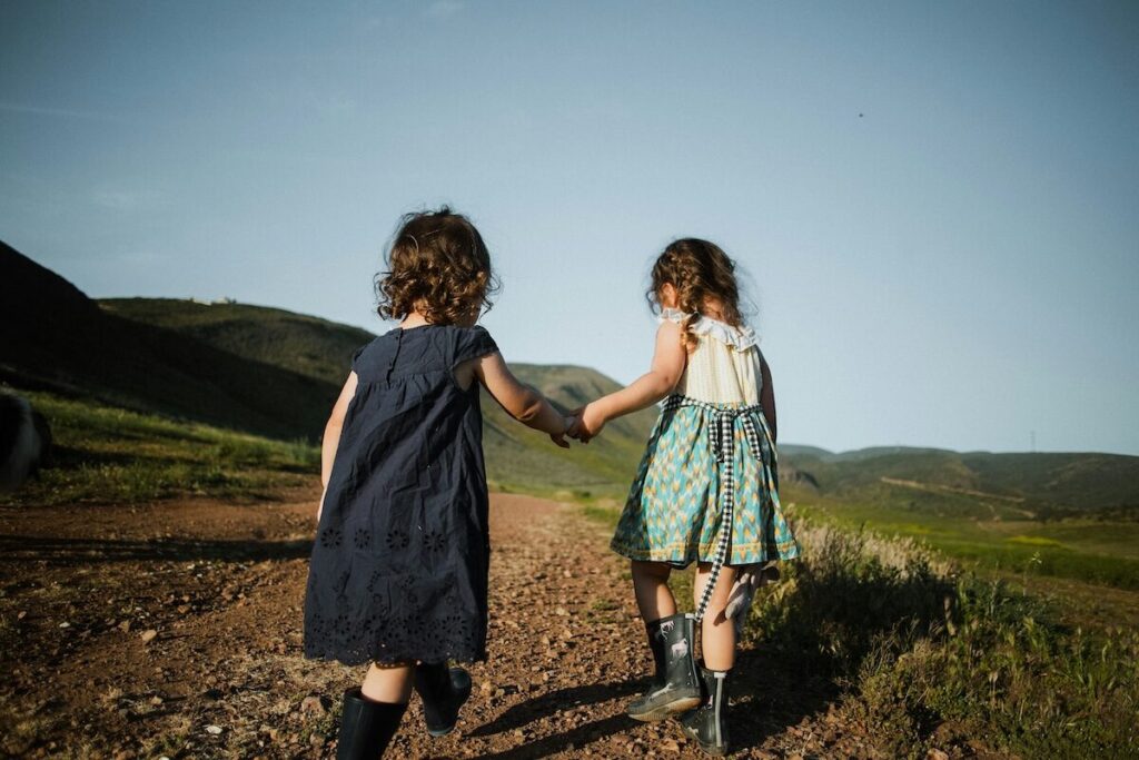Two young children walk hand in hand along a dirt path through a natural, grassy landscape with hills in the background.