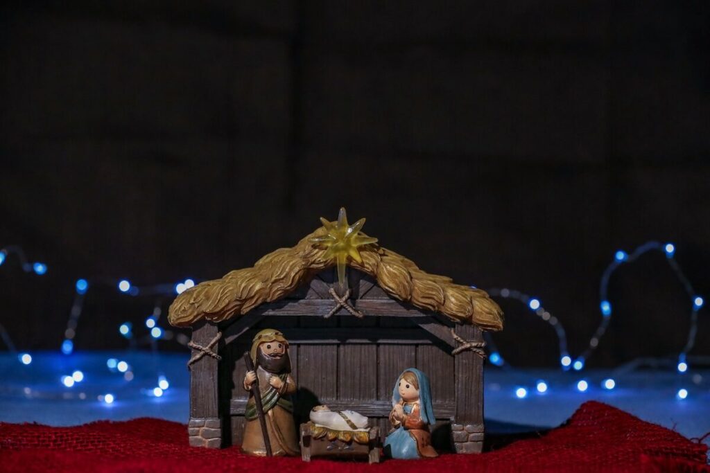 Nativity scene figurines of Mary, Joseph, and baby Jesus in a manger with a shed and decorative lights in the background.