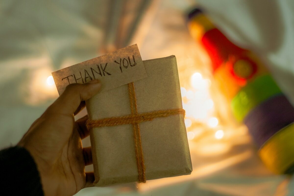 A hand holds a small wrapped gift with a Thank You note; blurred lights and a colorful object appear in the background.