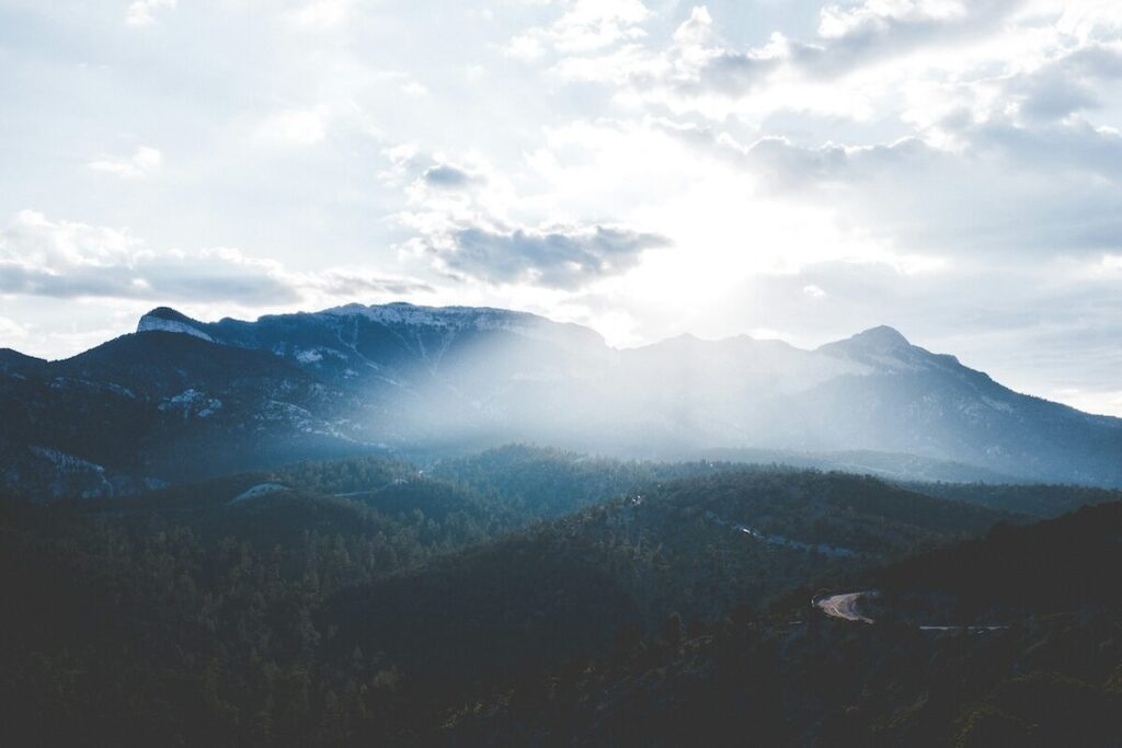 Sunlight streams through clouds over a range of forested mountains, creating a dramatic and serene landscape scene.