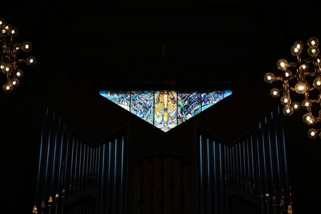 A triangular stained glass window above organ pipes, flanked by two chandeliers with round light bulbs, against a dark background.