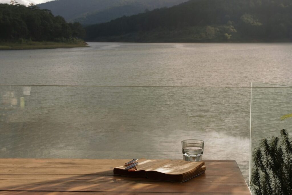 A wooden table with an open book, a pack of cigarettes, and a glass of water overlooks a calm lake with forested hills in the background.