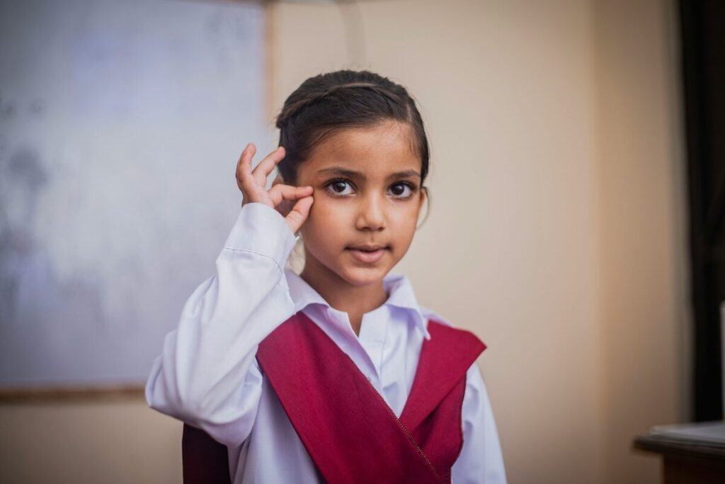 A young girl in a school uniform stands indoors, making a gesture with her hand while looking at the camera.