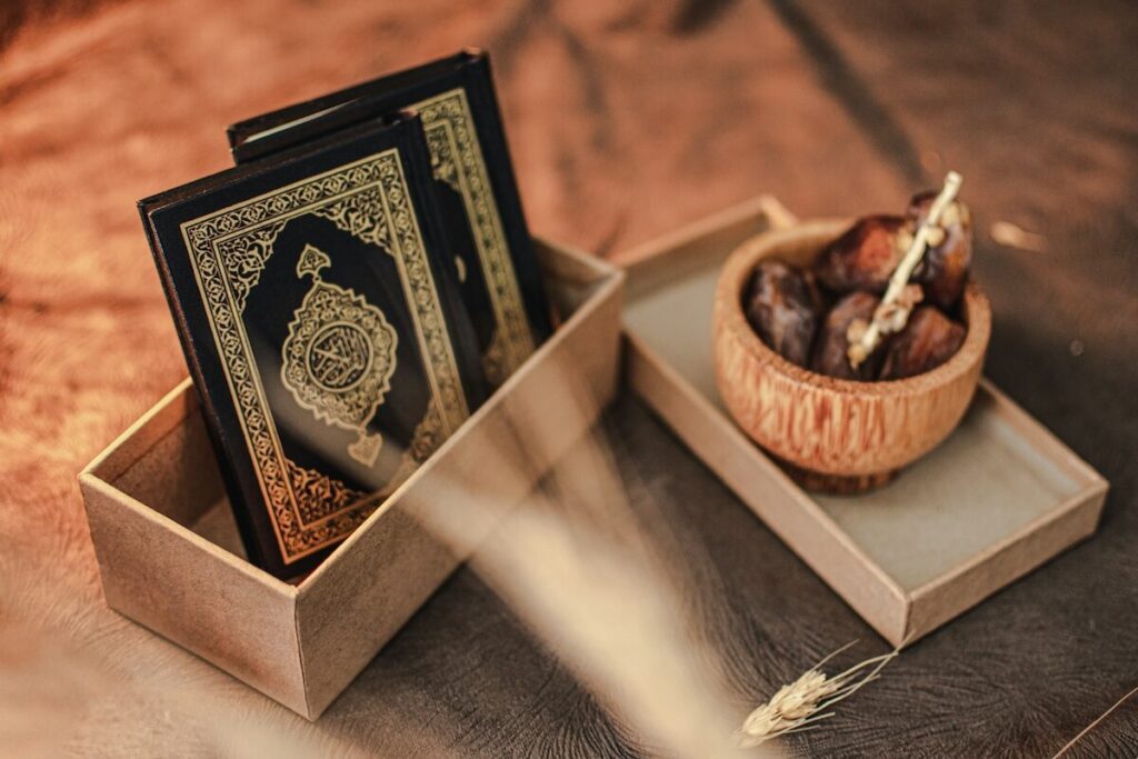 Two copies of the Quran in a box next to a bowl of dates with a stick, placed on a brown surface.