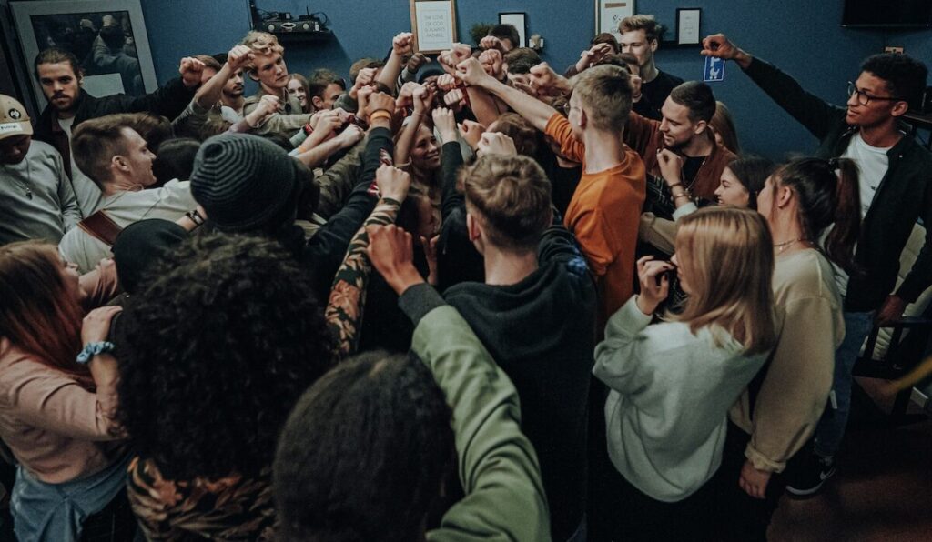 A large group of people stand in a circle indoors, raising their fists together in the center as a gesture of unity.