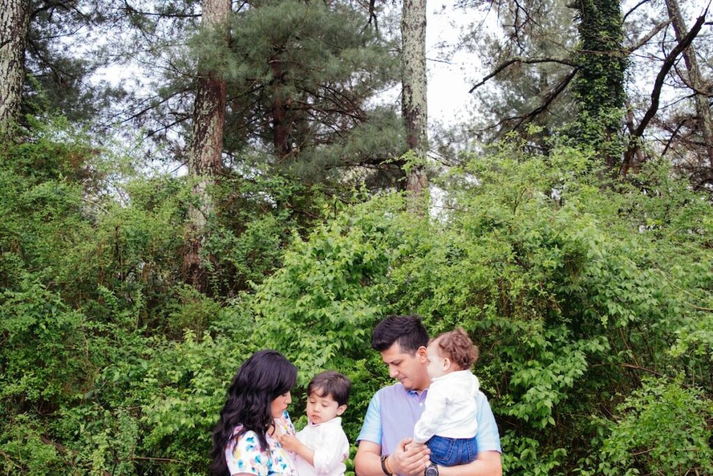 A family of four stands together outdoors in front of green bushes and tall trees, with two adults each holding a young child.