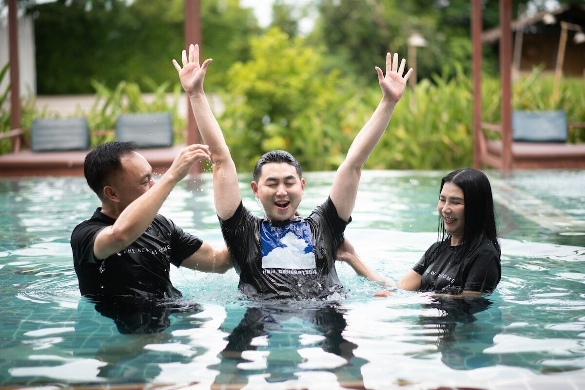 Three people stand in a pool, with the person in the middle raising their arms and smiling while the other two support them.