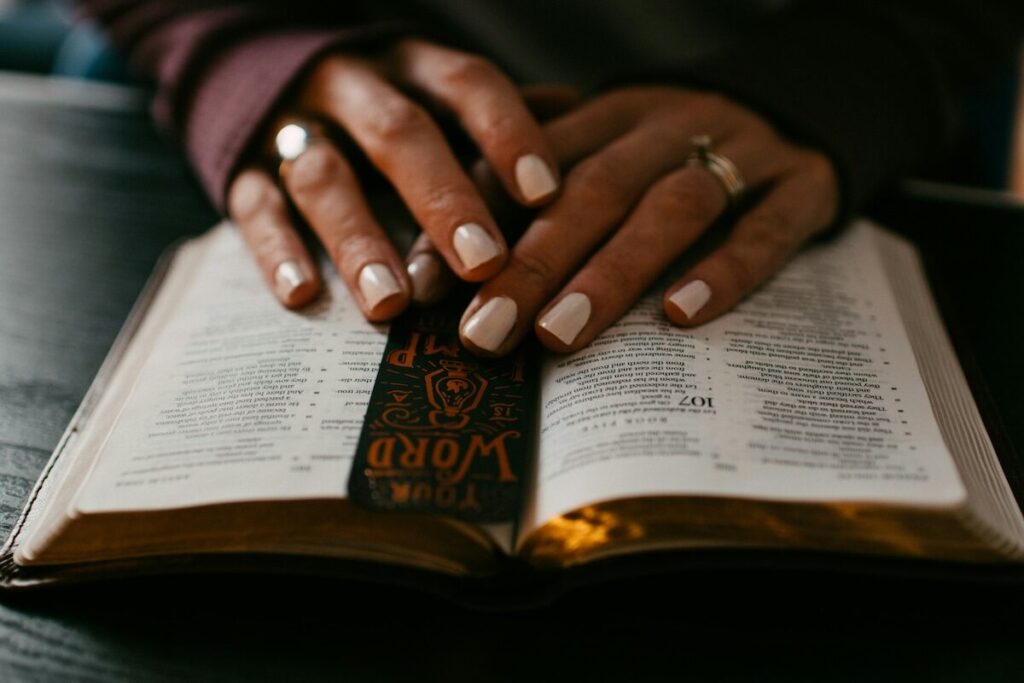 Close-up of hands resting on an open book, possibly a Bible, with a decorative bookmark placed inside.
