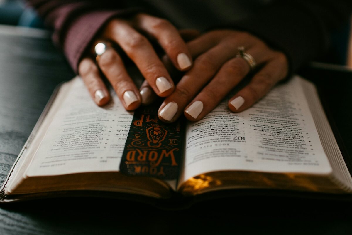 Close-up of hands resting on an open book, possibly a Bible, with a decorative bookmark placed inside.