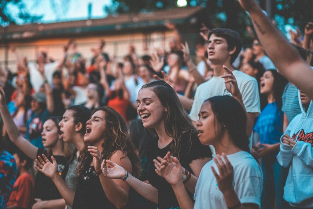 A large group of young people standing outdoors, raising their hands and singing or cheering together, with a blurred background.