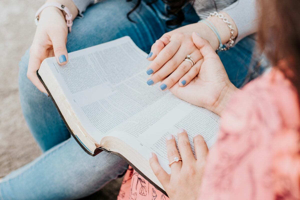 Two people sit closely together, holding and reading an open book, with one person’s hand resting on the page.