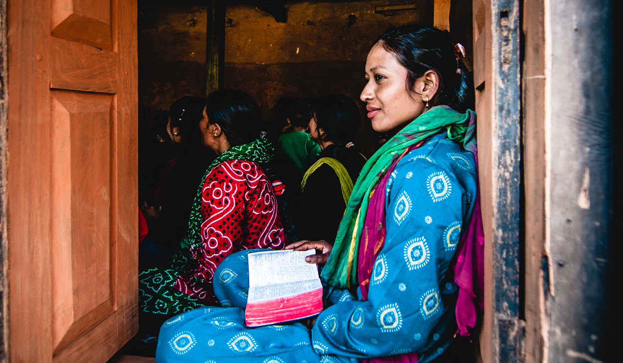 A woman in South Asia sits in a doorway with an open Bible. She is wearing a blue sari and a green scarf.