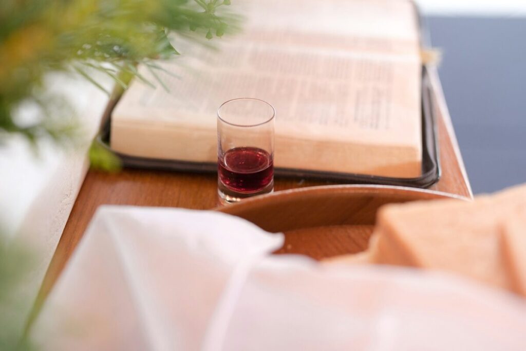 A small glass of red liquid sits on a wooden surface in front of an open book, with bread and a white cloth visible in the foreground.