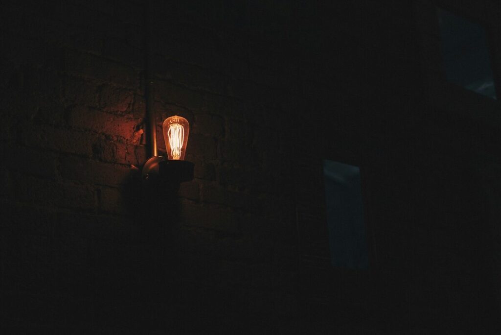 A single exposed light bulb mounted on a dark brick wall illuminates a small area, with most of the surroundings in shadow.