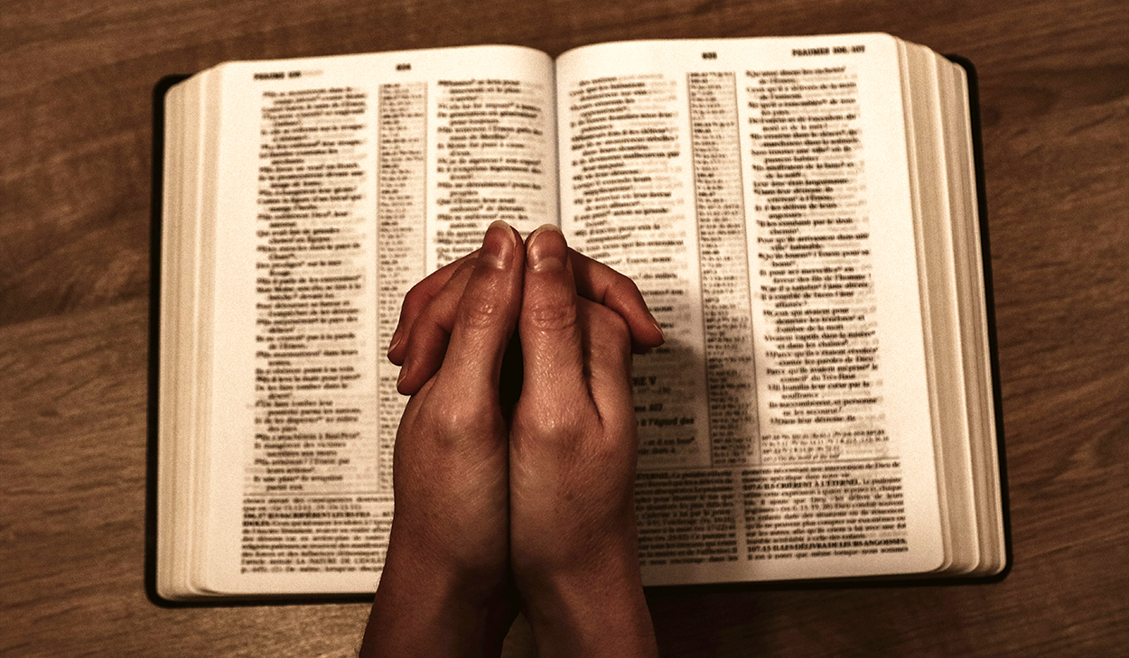 An individual folding their hands for prayer on top of an open Bible.