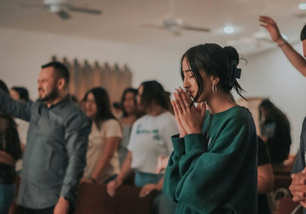 A group of people stand indoors, with a woman in the foreground clasping her hands together, appearing to be in prayer or deep thought.