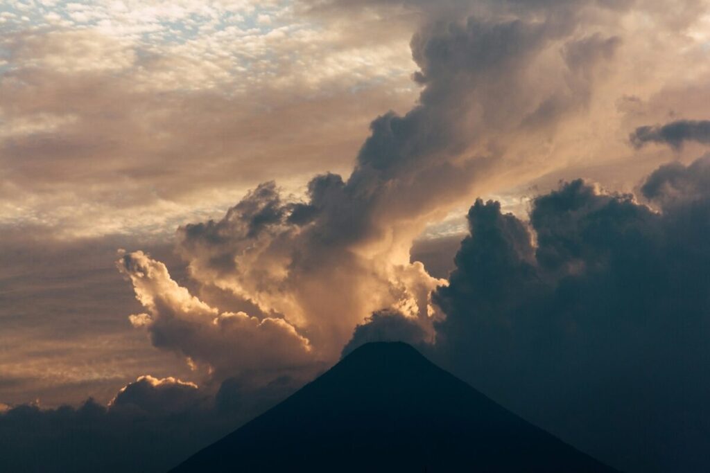 A dark silhouette of a mountain peak is set against dramatic clouds and a sky tinged with warm light during either sunrise or sunset.