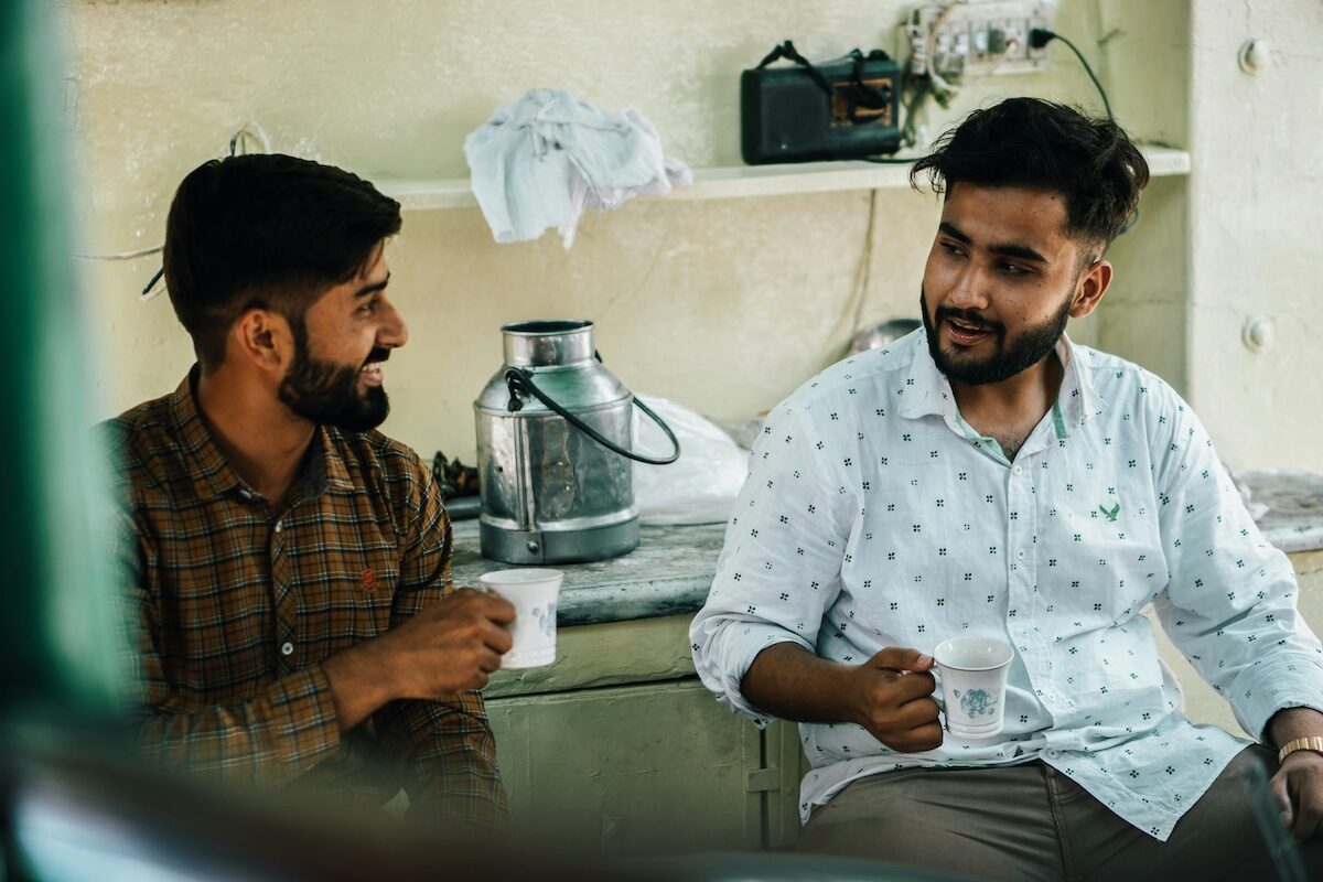 Two men sit indoors, holding mugs and having a conversation. A metal milk container and other items are visible on a shelf behind them.