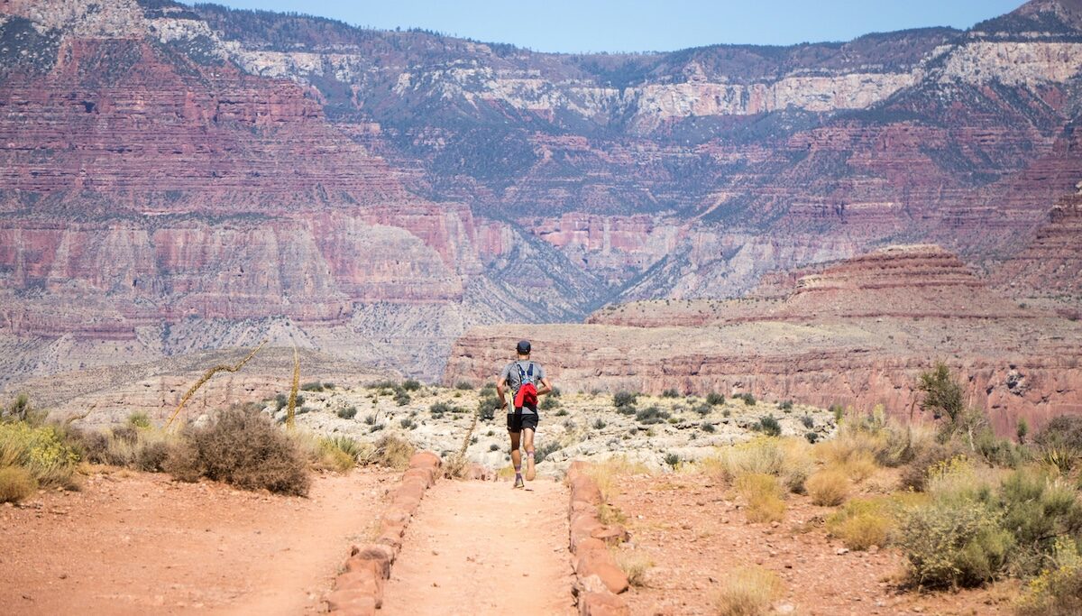 A person with a backpack walks along a trail through a dry, rocky landscape with layered red and tan canyon walls in the distance.