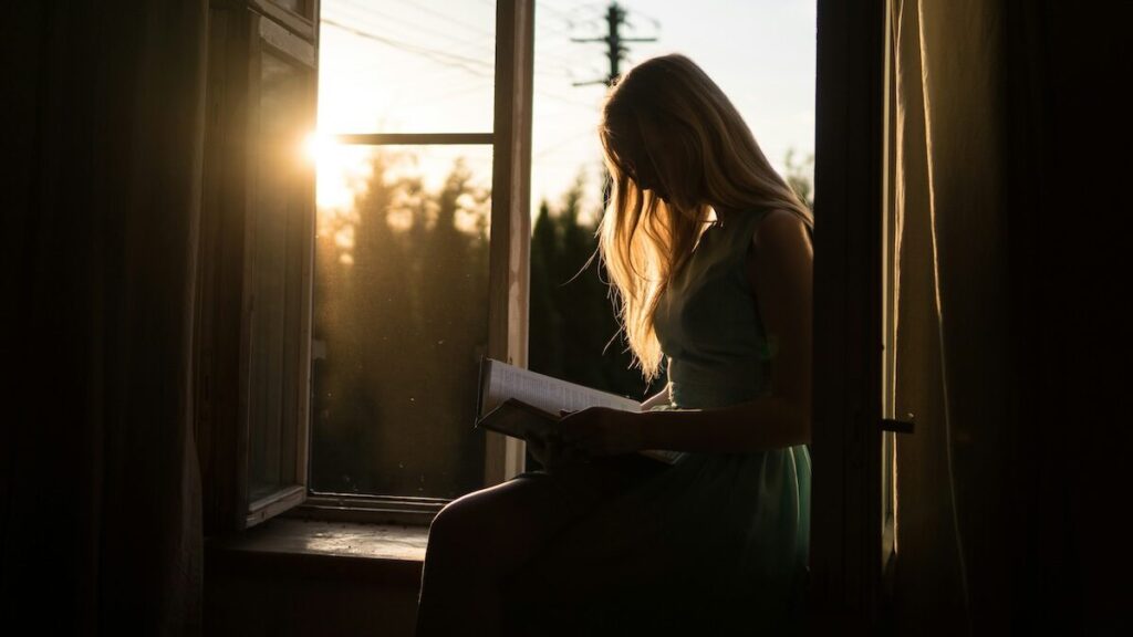 A person sits on a windowsill reading a book, with sunlight streaming through the open window in the background.