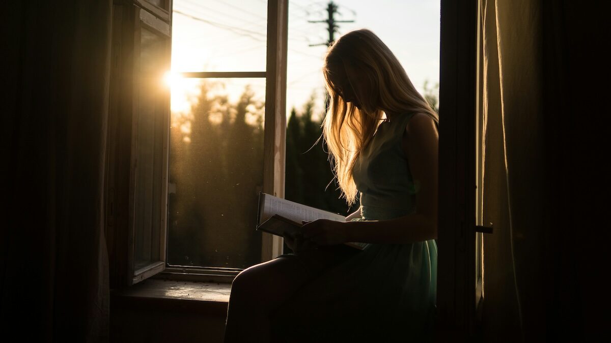 A person sits on a windowsill reading a book, with sunlight streaming through the open window in the background.