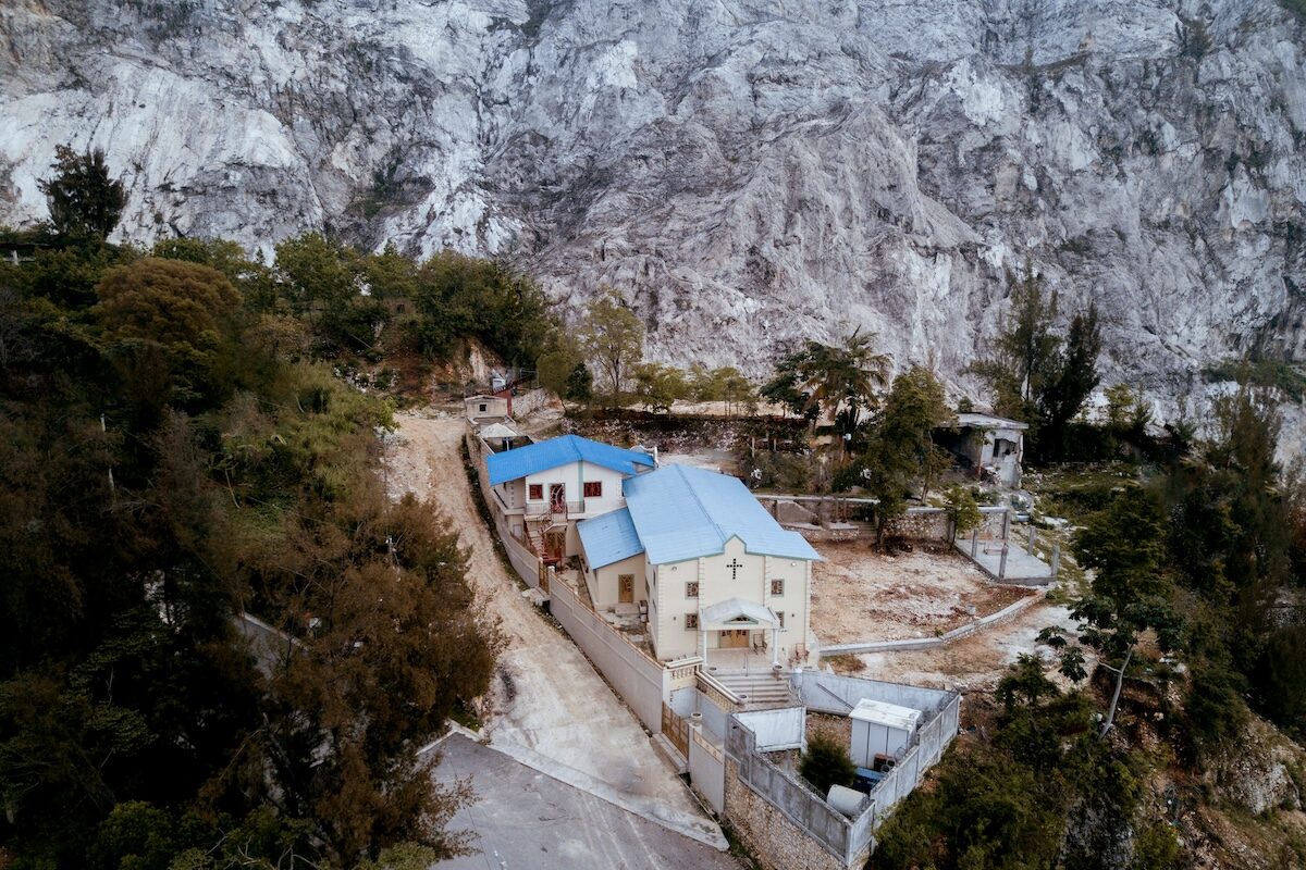 A small building complex with blue roofs is nestled among trees at the base of a rocky, mountainous landscape.