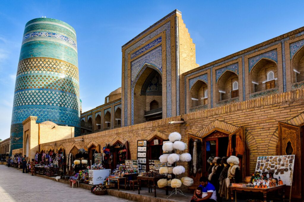A historic building with patterned brickwork and a large turquoise-tiled minaret, with market stalls selling goods in front.