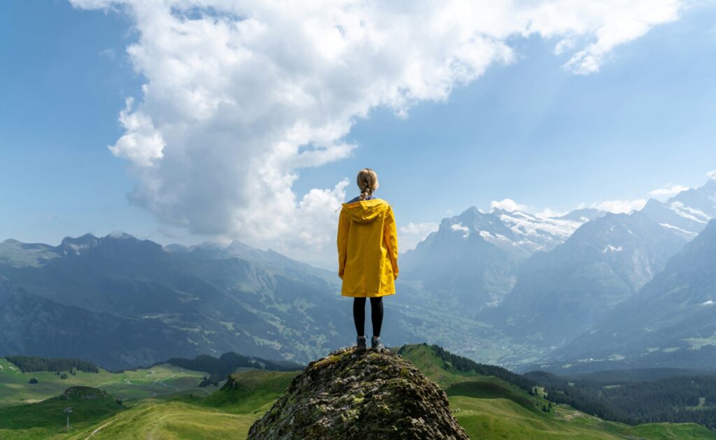 Person in a yellow jacket stands on a rock overlooking a vast valley with mountains and clouds in the distance.