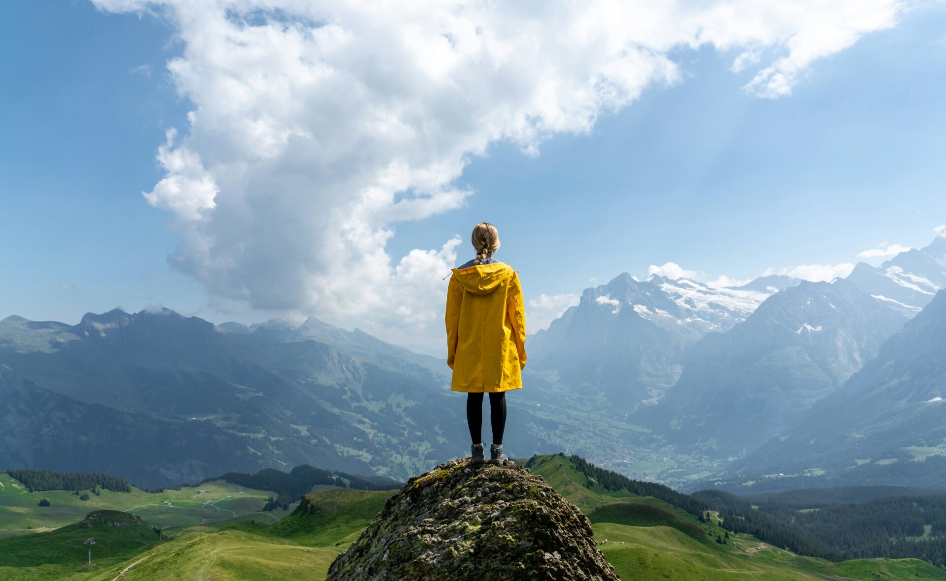 Person in a yellow jacket stands on a rock overlooking a vast valley with mountains and clouds in the distance.