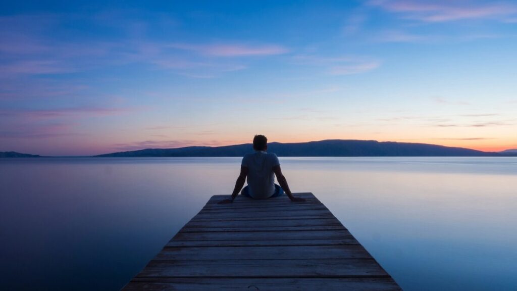 A person sits at the end of a wooden dock overlooking calm water and distant hills during sunset or sunrise.