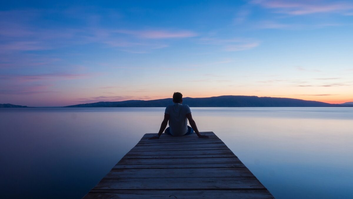 A person sits at the end of a wooden dock overlooking calm water and distant hills during sunset or sunrise.