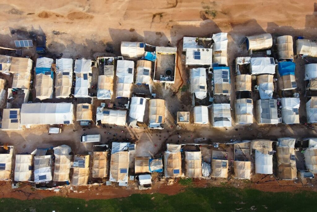 Aerial view of a refugee camp with rows of makeshift shelters and tents, surrounded by bare earth and patches of greenery.