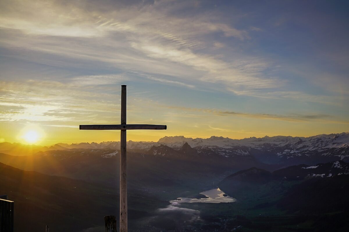 A wooden cross stands on a hilltop with a sunrise over mountains and a lake in the background.