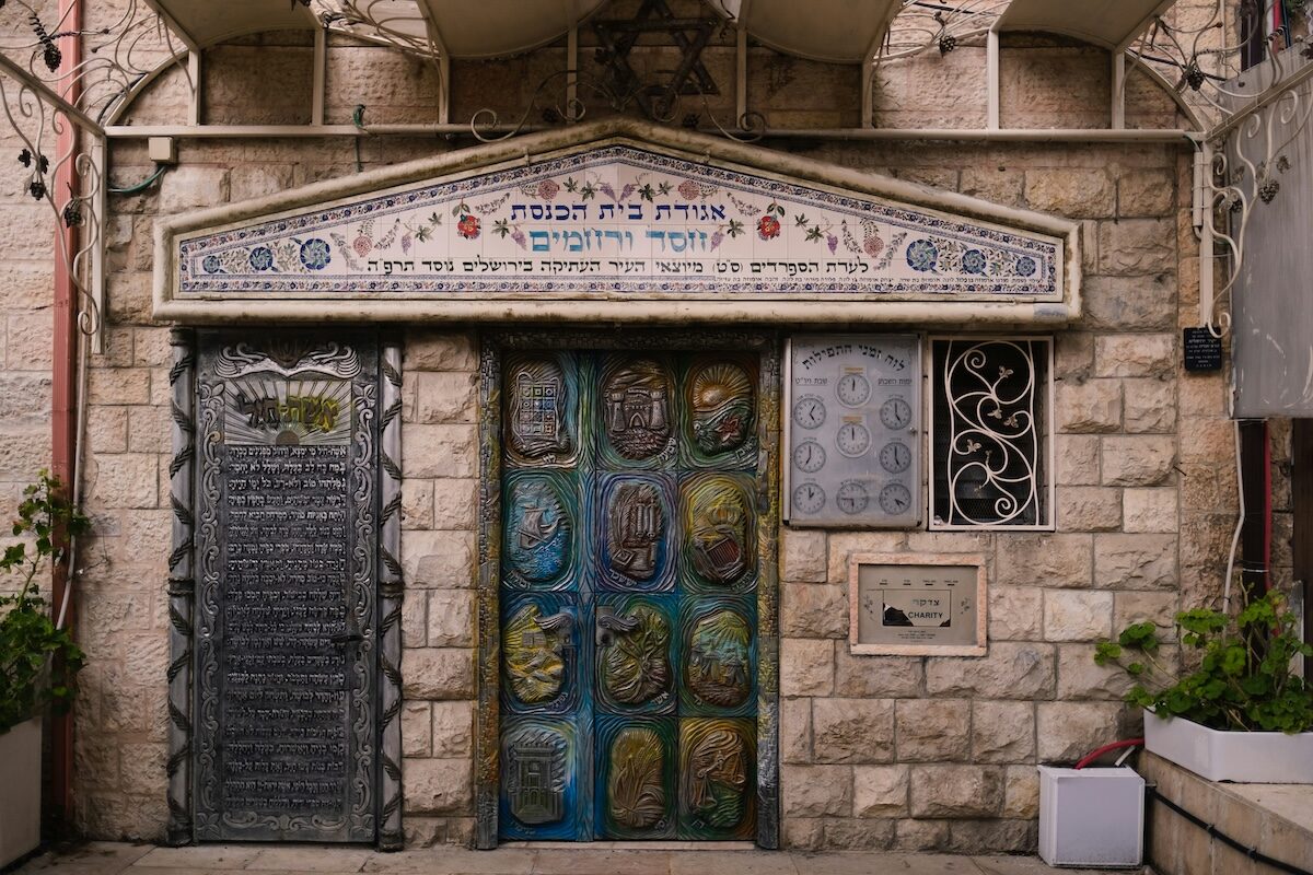 Stone building façade with Hebrew inscriptions above a decorated metal door, a black plaque on the left, and a small window with a metal grille on the right.