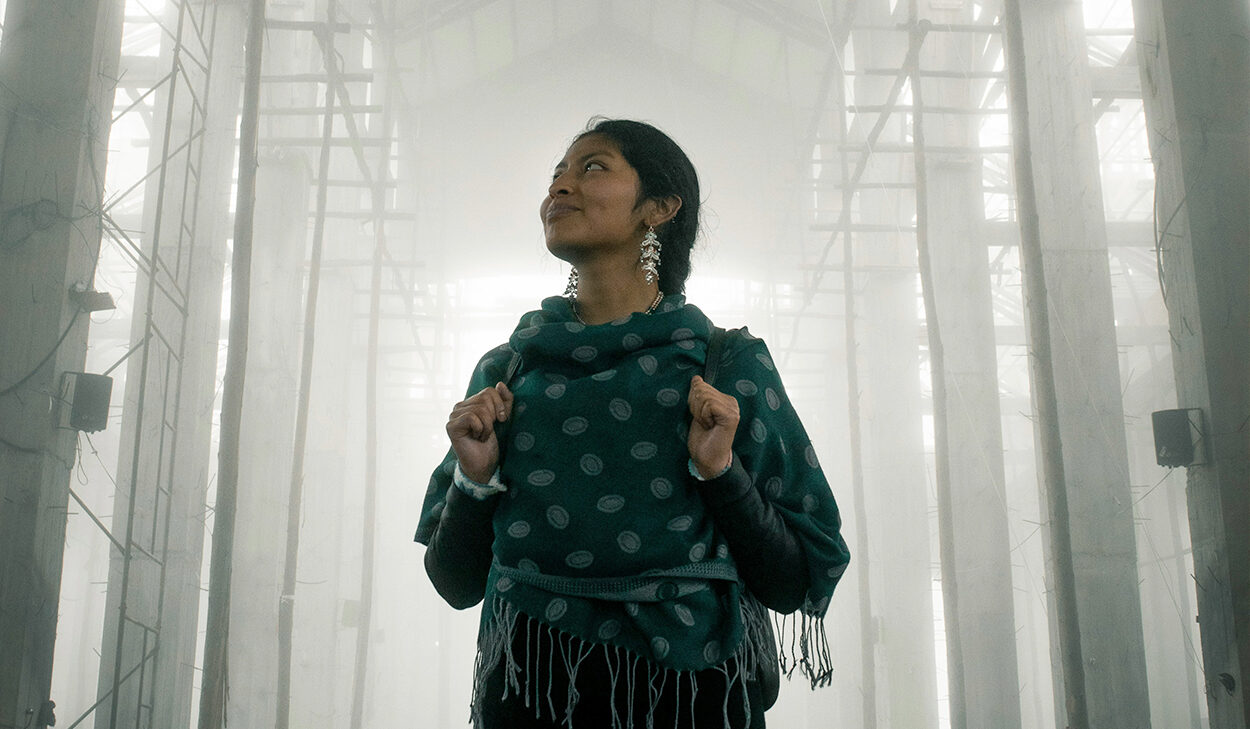 A woman with her dark hair in a braid stands in the middle of a well-lit sanctuary, looking up to the ceiling.