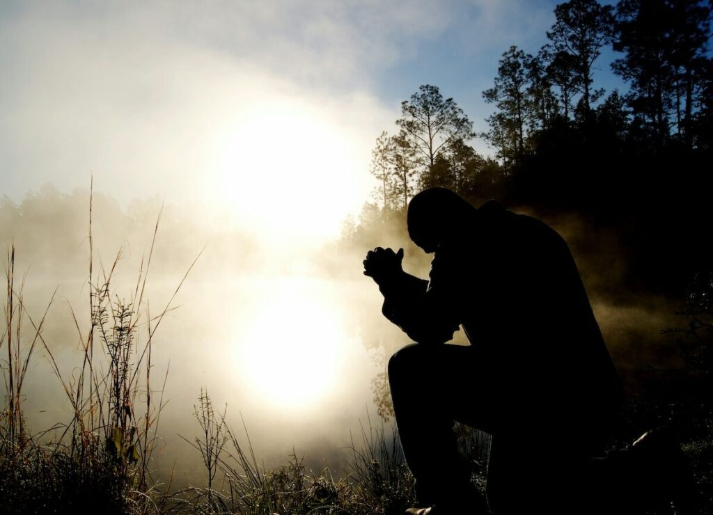 A person kneels outdoors at sunrise or sunset, silhouetted against a bright sky with trees and tall grasses in the background.
