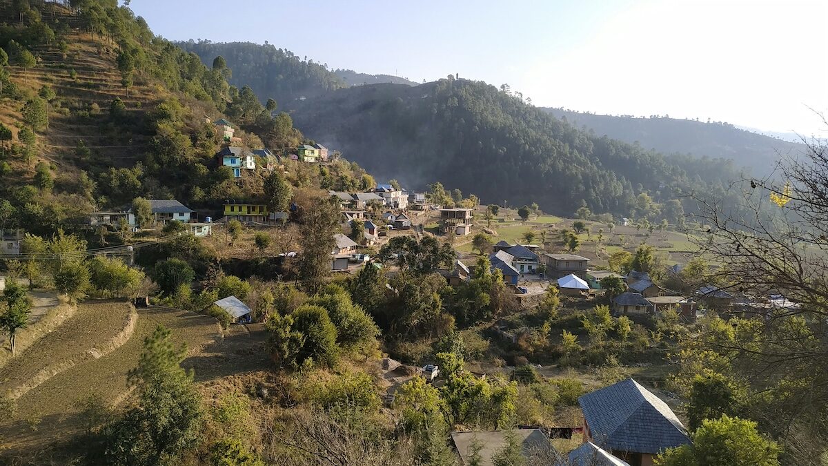 A small village with scattered houses sits in a green hilly landscape, surrounded by trees and terraced fields under a clear sky