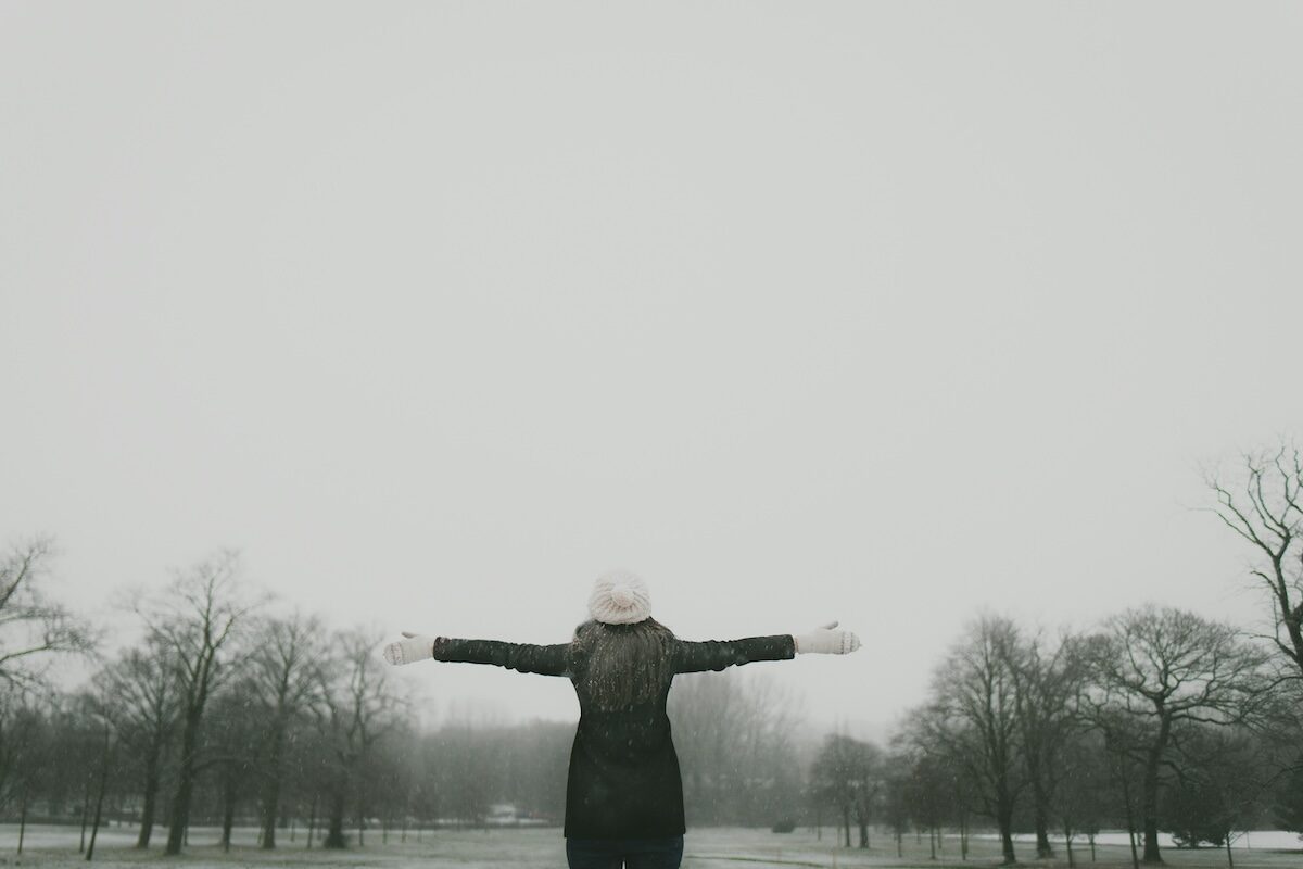 A person stands with arms outstretched in an open, foggy park surrounded by leafless trees.