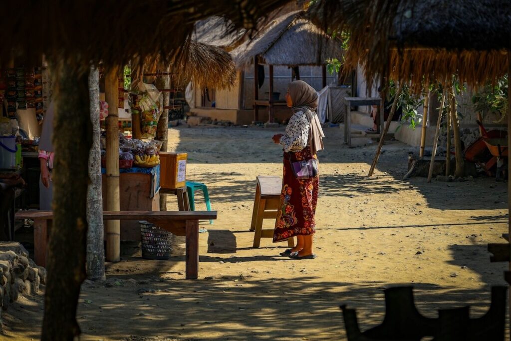 A woman wearing a headscarf and patterned skirt stands in a sandy outdoor area surrounded by thatched structures and wooden benches.