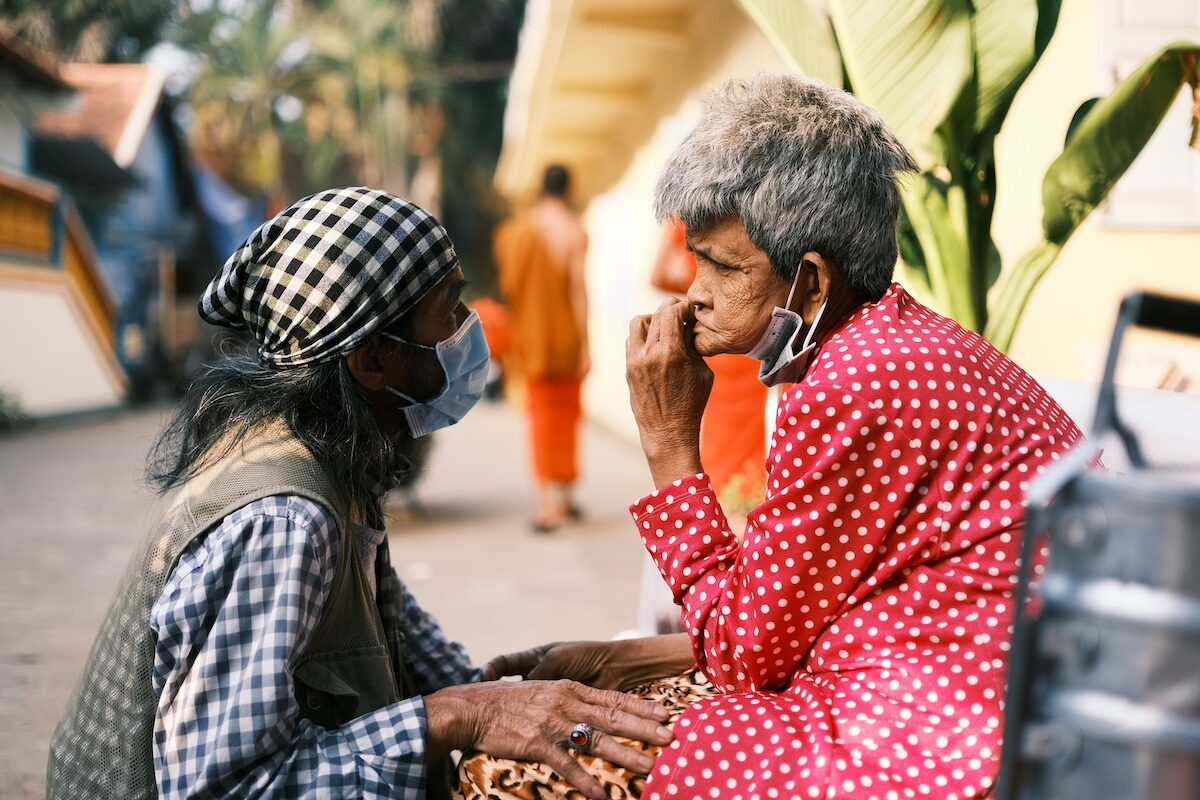Two elderly people wearing face masks sit close together outdoors, holding hands and engaging in conversation. This caring image captures the warmth of companionship, with one in a checkered headscarf and the other in a red polka-dot outfit.