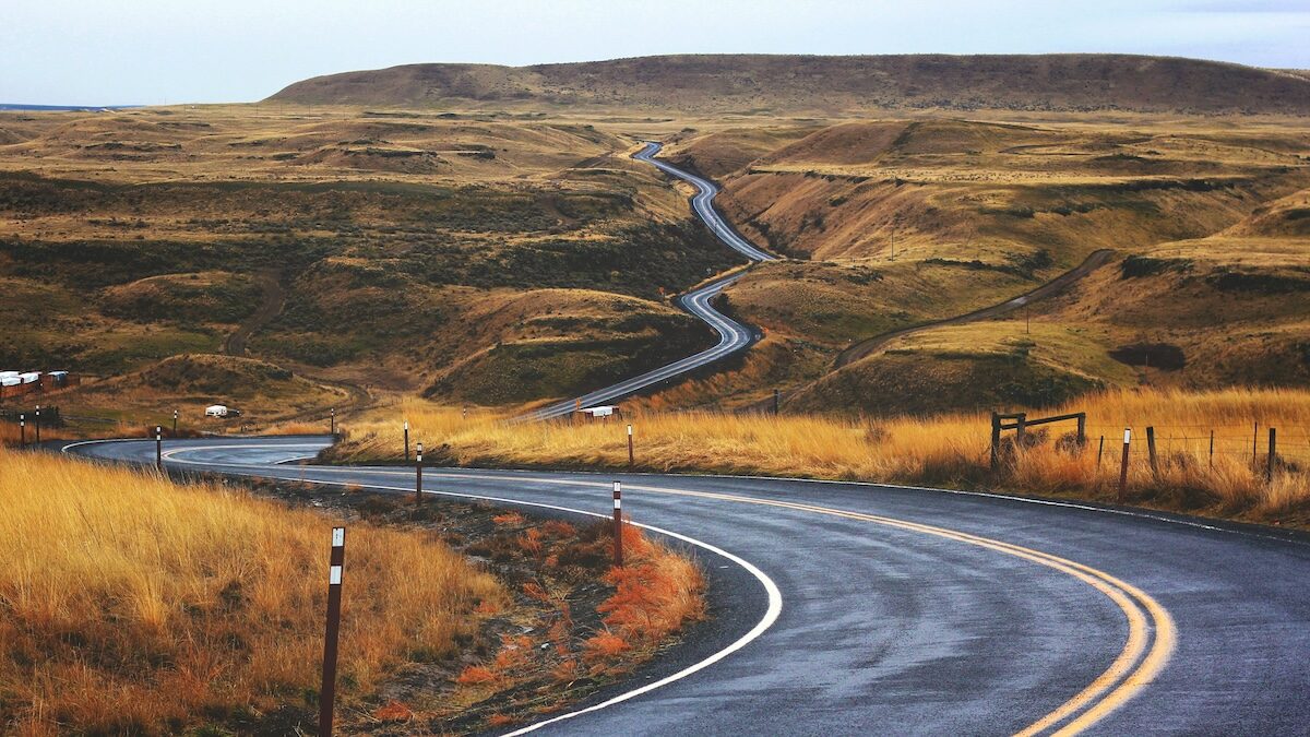 A winding paved road curves through a hilly, grassy landscape under a cloudy sky.