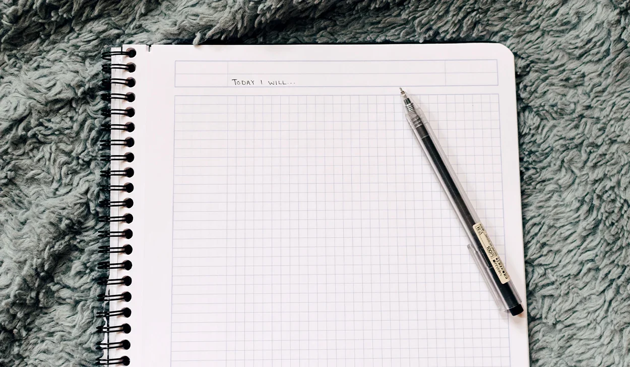 An open spiral notebook with graph paper rests on a textured blanket. The top line reads Today I will.... A black pen is placed on the right side of the notebook.