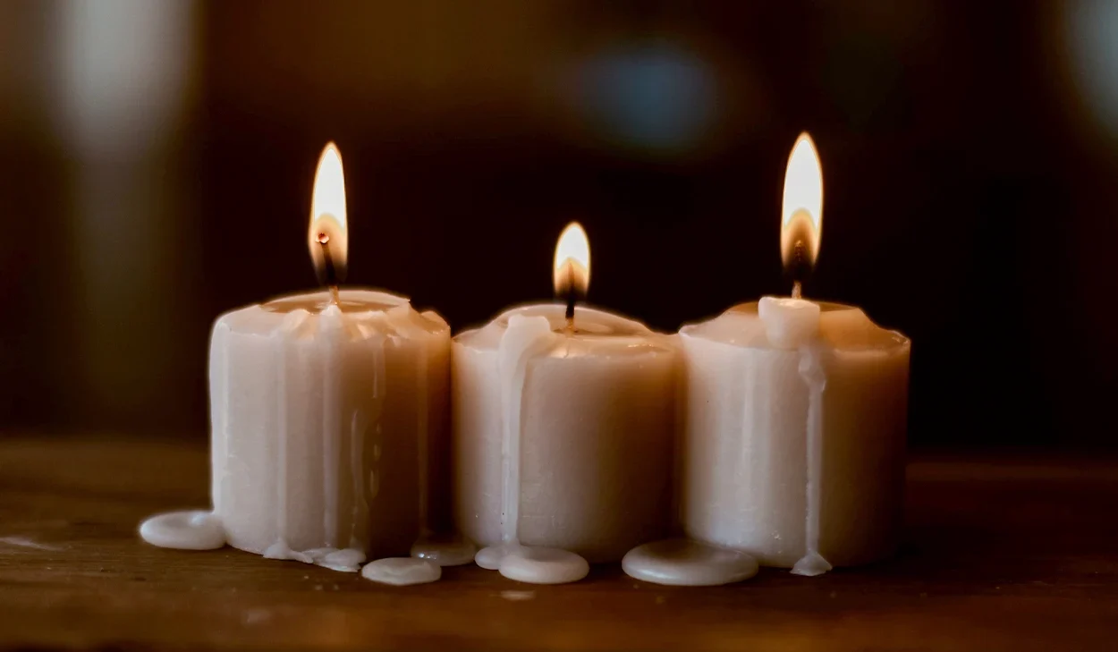 Three white candles with melted wax are burning on a wooden surface, each with a visible flame and wax dripping down the sides.
