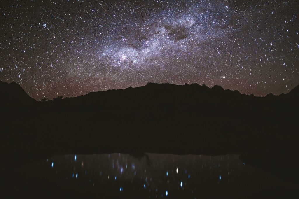 The silhouette of mountains under a night sky filled with stars, with their reflections visible in a dark, still body of water below.