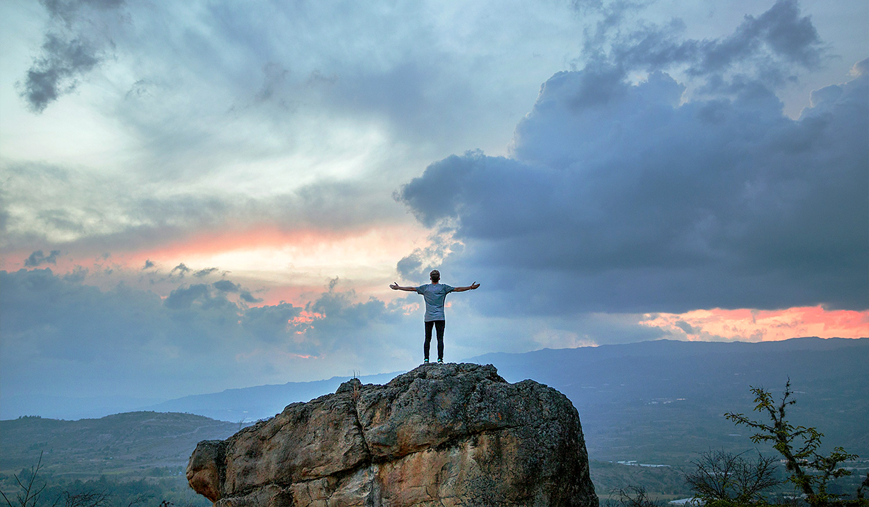 A man in a white t-shirt stands on top of a boulder with his arms outstretched. He is facing a valley as the sun rises among the clouds.